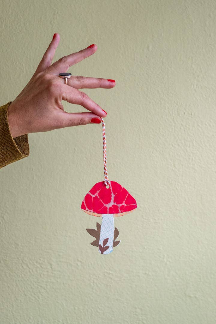 A hand with red nail polish holds a small red and white paper cutout mushroom ornament with a red-and-white string, set against a textured beige wall.
