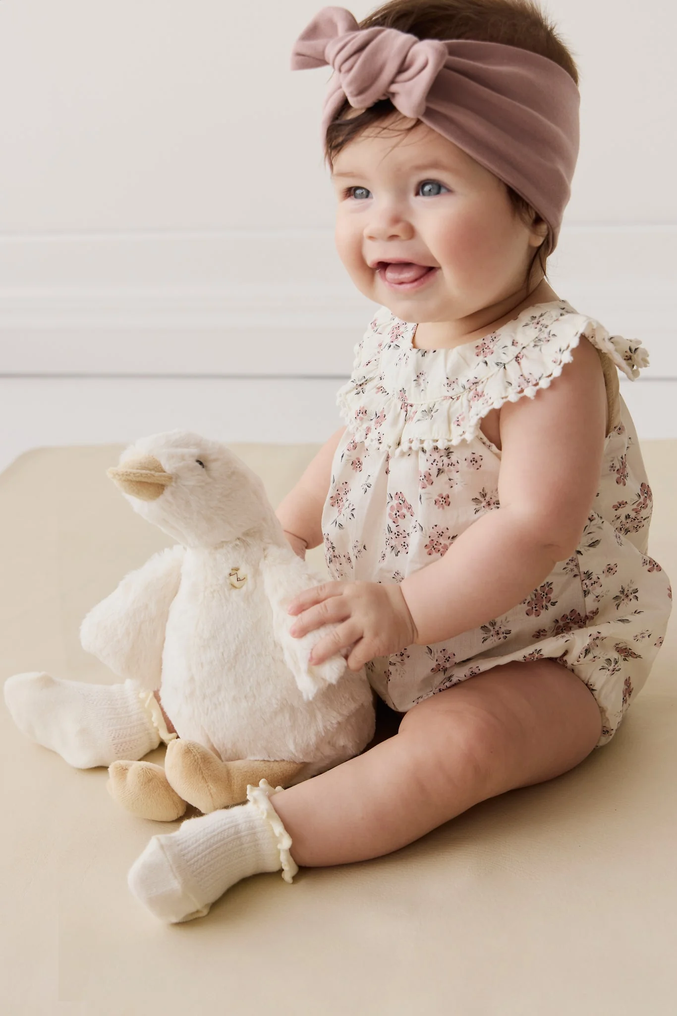 Baby girl in floral outfit with teddy bear on beige surface