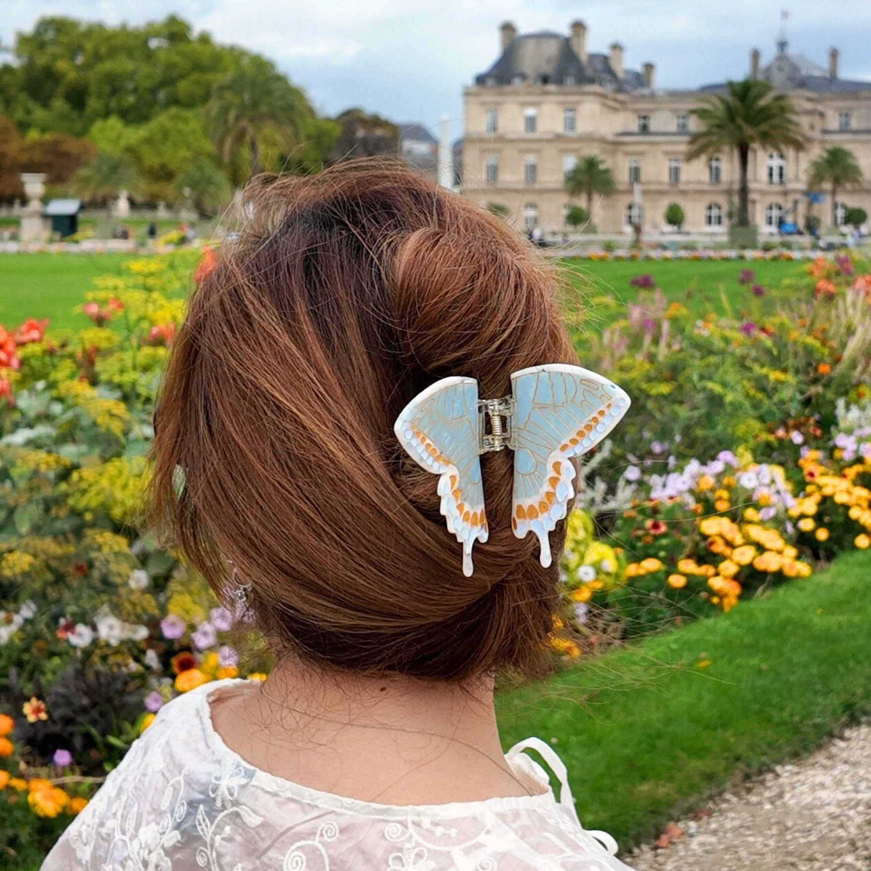 Person sitting in a garden with a butterfly hair clip, looking at a large building.