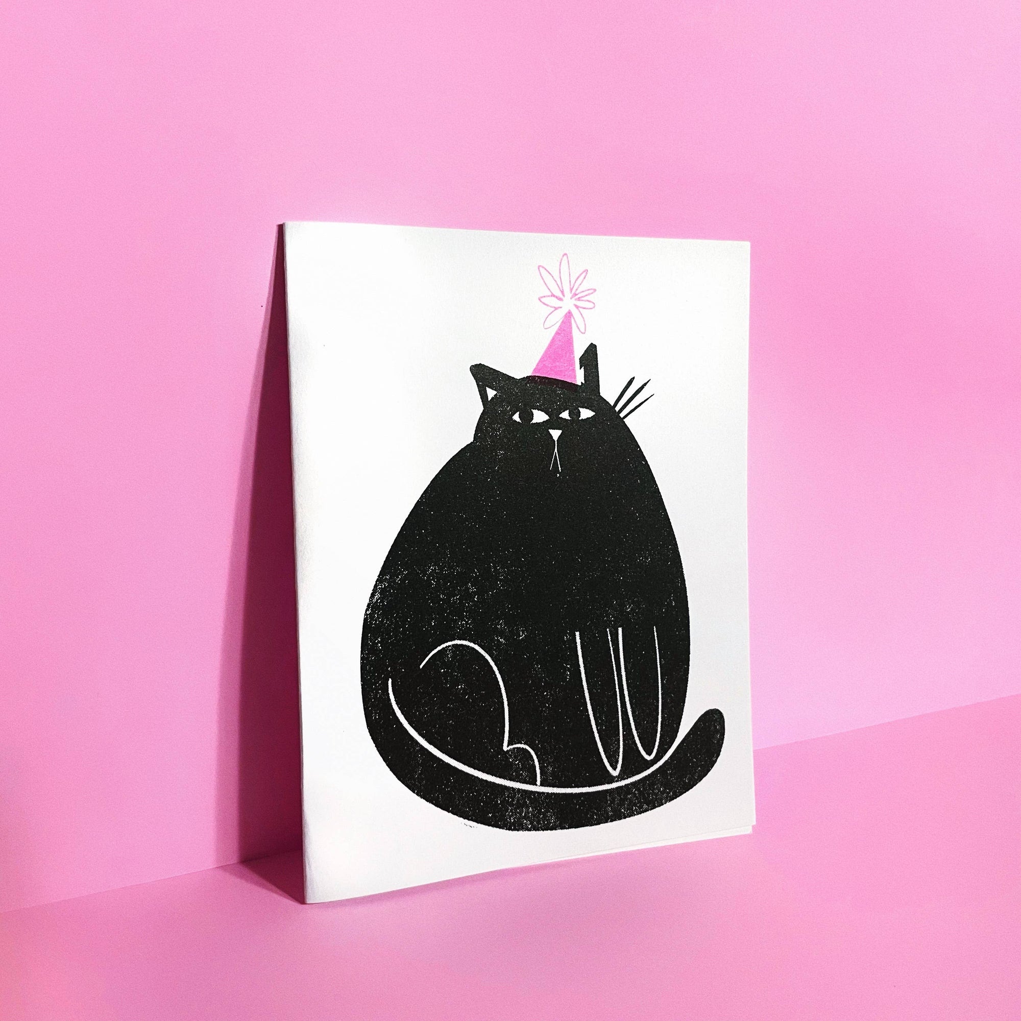 Greeting card with a black cat wearing a pink party hat on a pink background