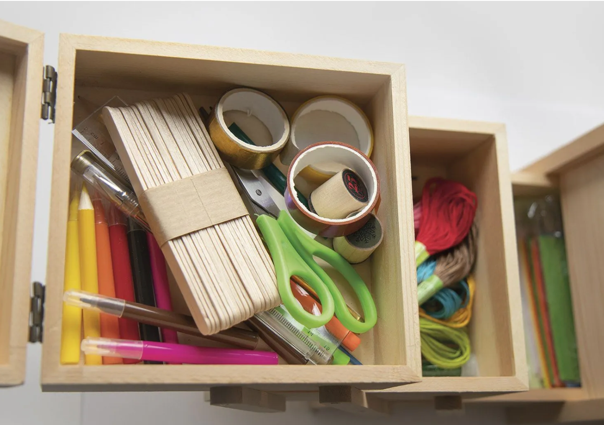 An open wooden organizer drawer filled with colorful stationery and craft supplies, including pencils, markers, scissors, tape rolls, and thread balls, with another drawer partially visible beside it.