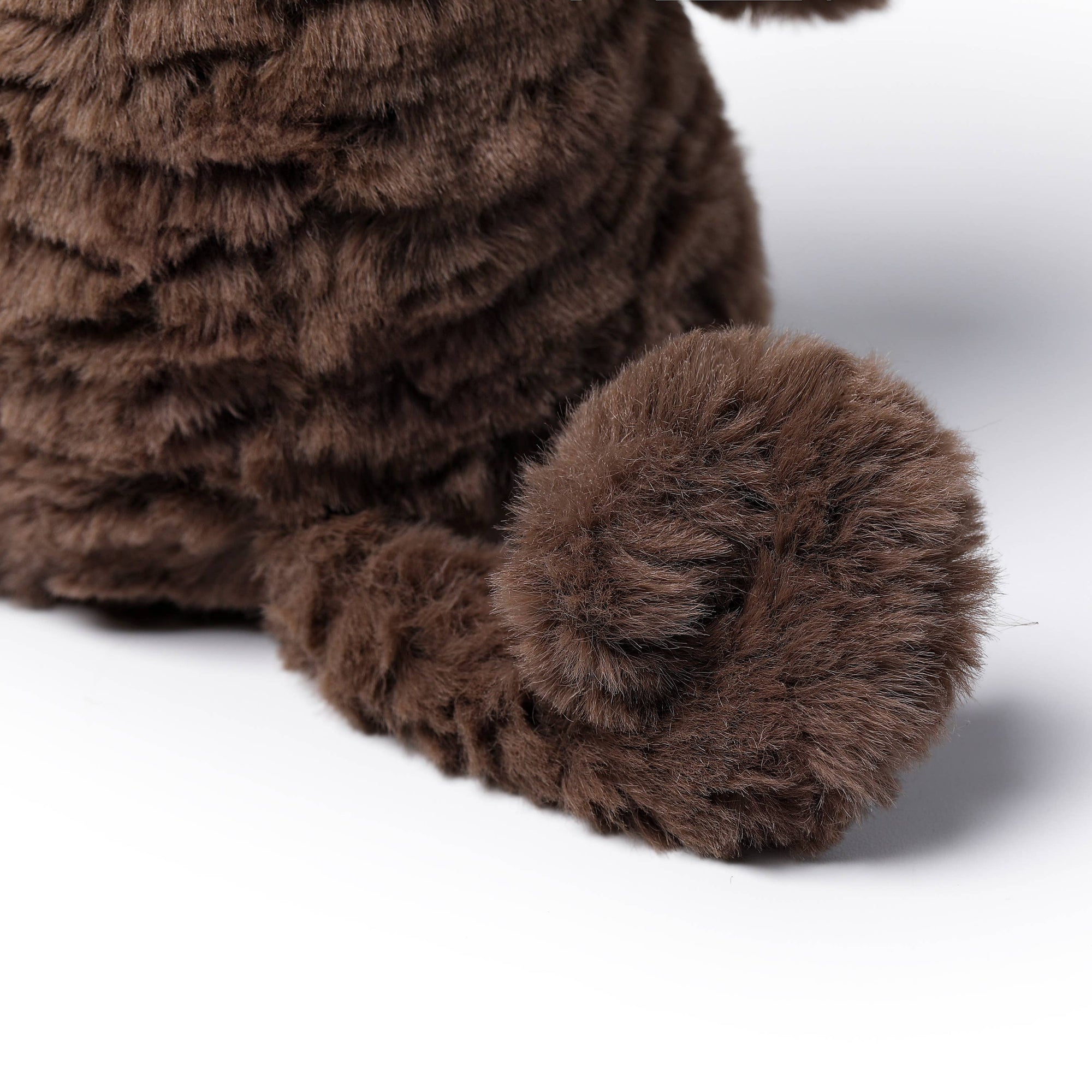Close-up of a brown plush toy with a shaggy, textured fur-like surface and a curled tail, photographed on a clean white background.