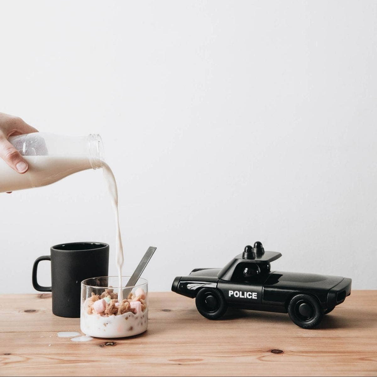 Person pouring milk into a glass with cereal next to a toy police car on a wooden table.