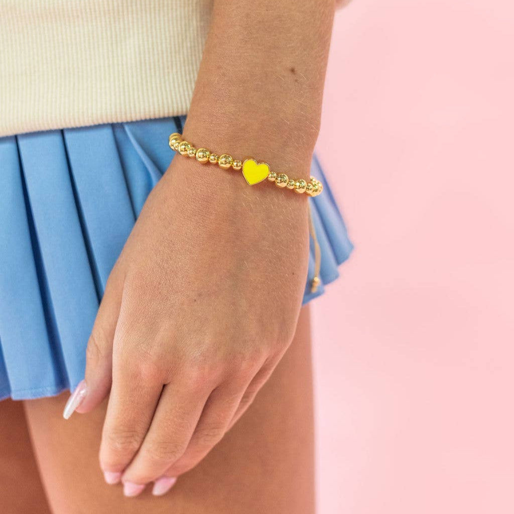 Hand wearing a gold bracelet with a yellow heart charm on a pink background