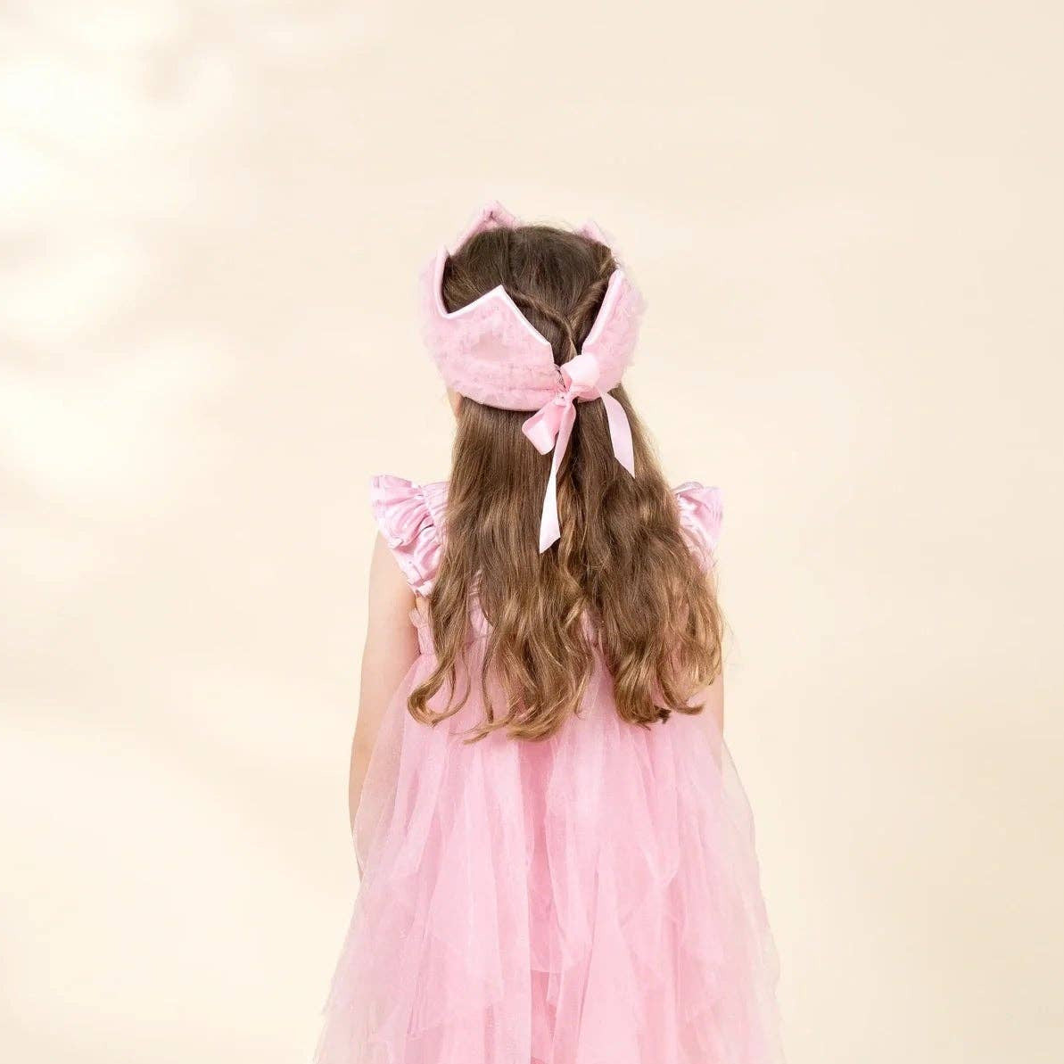 A young girl viewed from behind wearing a pink tiered tulle dress, a matching pink bow headband, and pink ballet slippers, standing against a warm cream background.