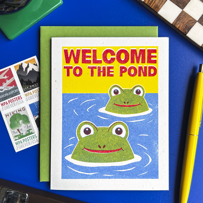 A square greeting card with a green envelope, featuring a colorful illustration of two green frogs in blue water with the text 'WELCOME TO THE POND' in red. The card is styled on a blue surface with vintage WPA posters, a yellow pen, and a green object in the background.
