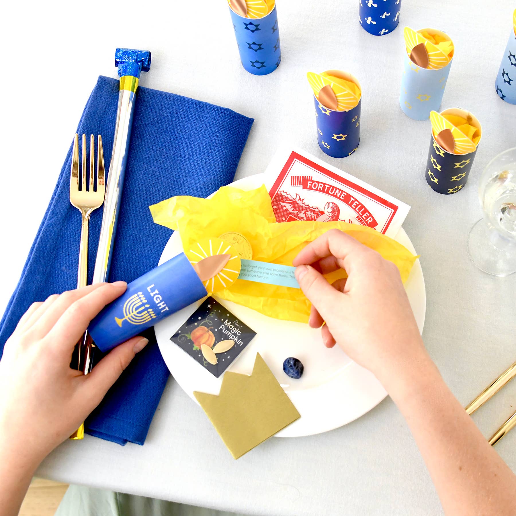 Hands setting a festive table with blue napkins, gold cutlery, and colorful candles.