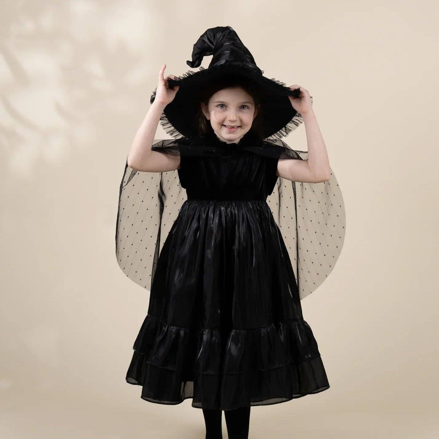 A young girl modeling a black witch costume dress with a matching pointed witch hat, sheer black polka-dot wings, and black tights against a warm beige studio background.
