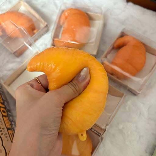 A hand holding a bright orange, crescent-shaped squishy toy, with several similar orange squishies in clear plastic packaging on a white textured surface in the background.
