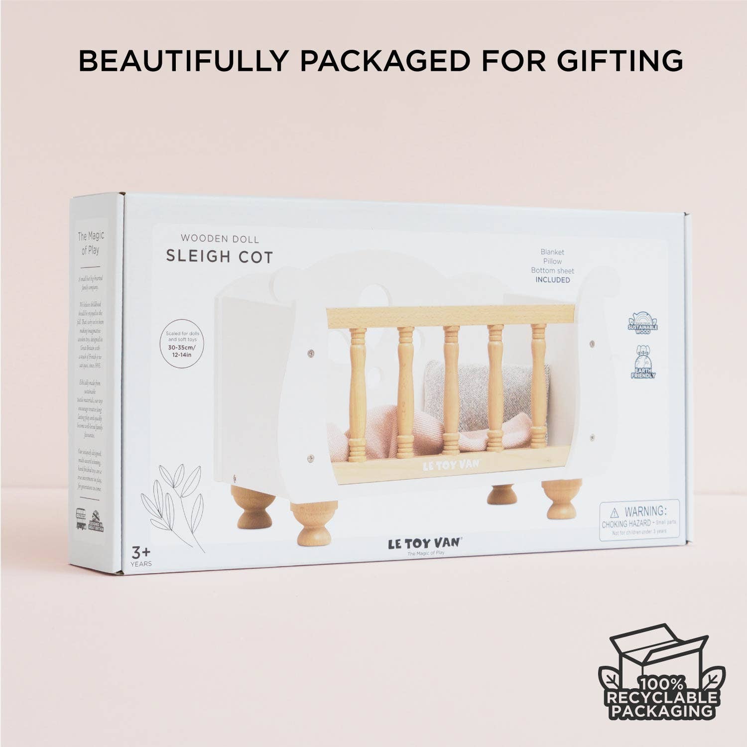 Packaged wooden doll sleigh cot with branding and recyclable packaging label on a light pink background