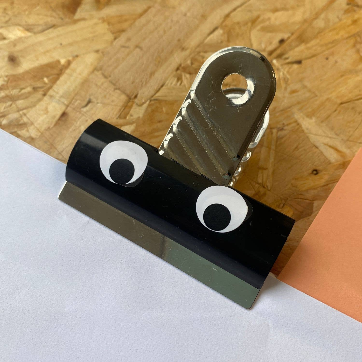 Black paper clip with googly eyes on a wooden surface