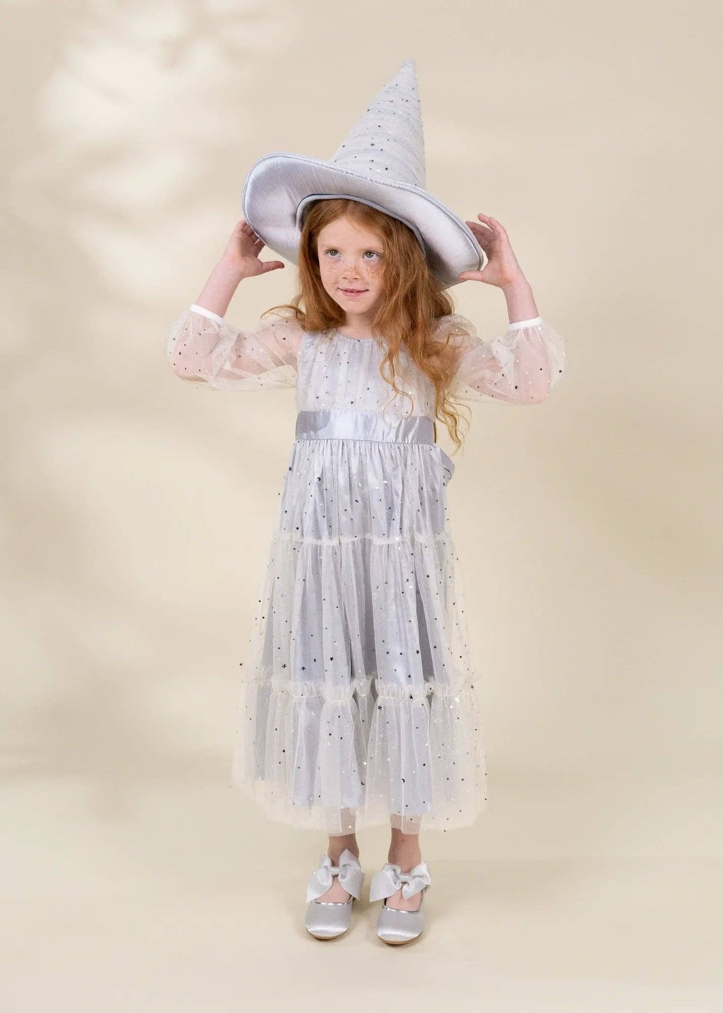 A young girl with long red hair stands against a warm beige studio background wearing a white wizard costume dress with silver glitter details, a pointed wizard hat, and silver shoes.
