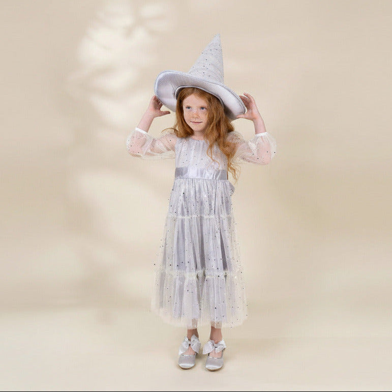 A young girl with long red hair stands against a warm beige studio background wearing a white wizard costume dress with silver glitter details, a pointed wizard hat, and silver shoes.