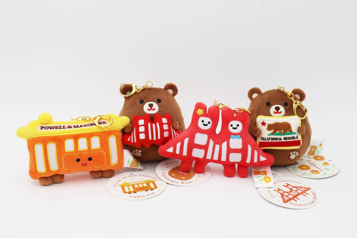Three small plush bear-shaped keychains or charms arranged on a white background, each with a printed food-themed label and a small gold key ring. The left figure is orange and yellow with a bus-like design and the text 'Powell & Mason'; the center figure is red and white with a taco design; the right figure is brown with a pizza design and the text 'California Roll'.