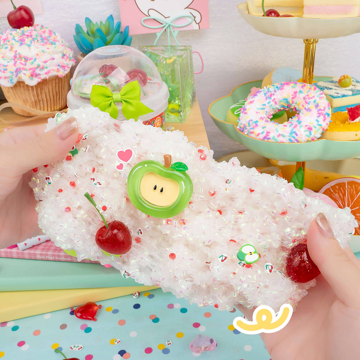A close-up of hands holding a white glittery headband decorated with a green apple charm, a cherry, and small colorful heart and leaf stickers, set against a pastel dessert-themed background.