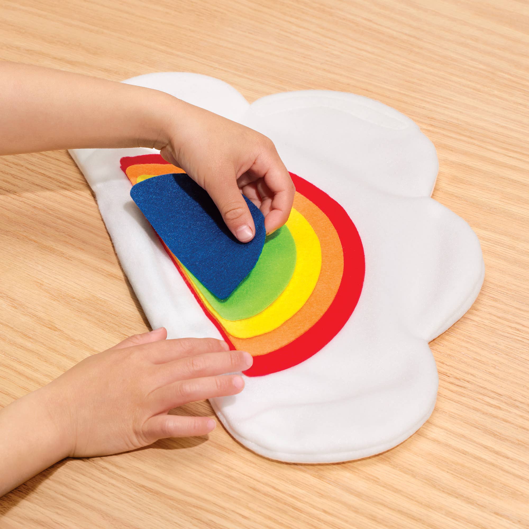 A child's hands are shown interacting with a white cloud-shaped felt mat on a light wood floor, lifting a colorful felt rainbow layer by layer.