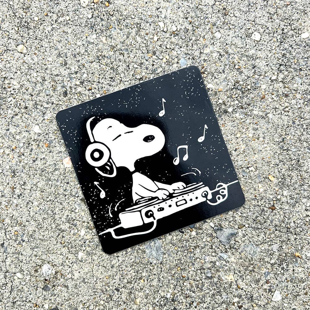 Sticker of a dog with headphones and DJ equipment on a black background, placed on concrete.