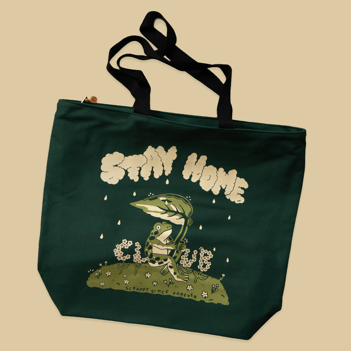 A dark green canvas tote bag with black handles, featuring a graphic print of a frog sitting on a hill with the text 'STAY HOME CLUBE' and 'SCRAPBOOK CLUBE' on a beige background.
