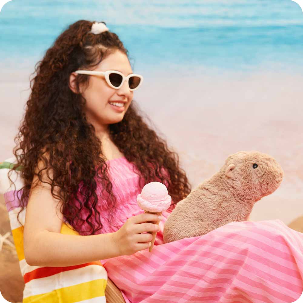 Woman in a pink dress with sunglasses holding an ice cream, sitting next to a plush capybara on a beach.