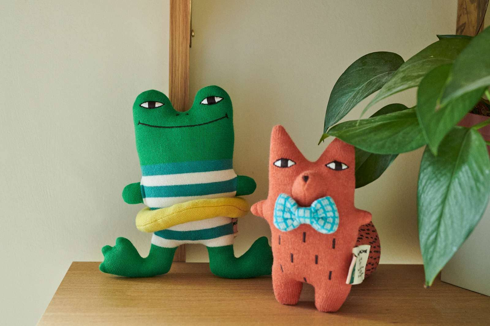 Two handmade plush toys on a wooden shelf: a green frog wearing a teal and white striped shirt with a yellow belly, and a pinkish-orange cat wearing a blue patterned bow tie, next to a green houseplant.
