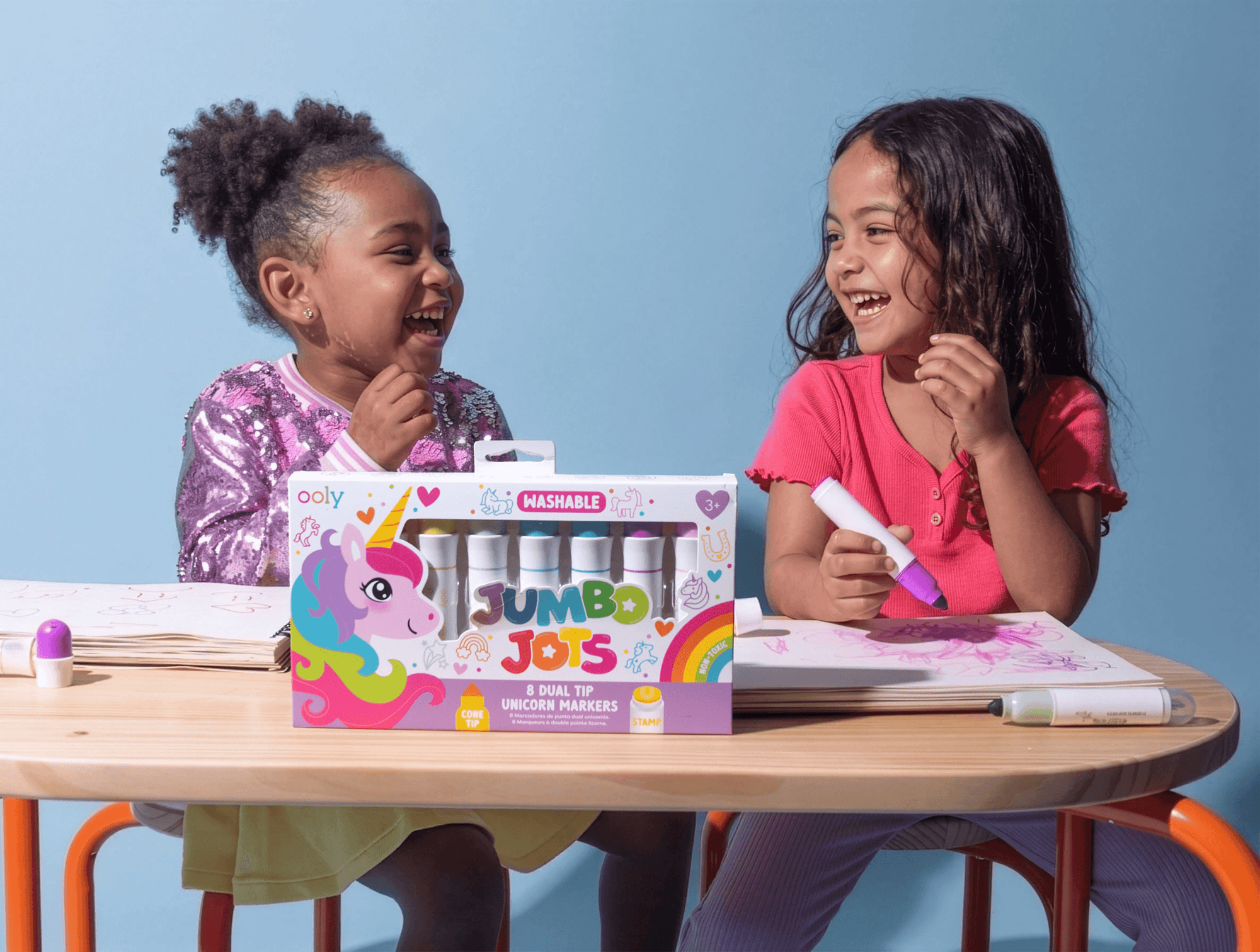 Two children sitting at a table with a box of Ody Jumbo Joys markers against a blue background