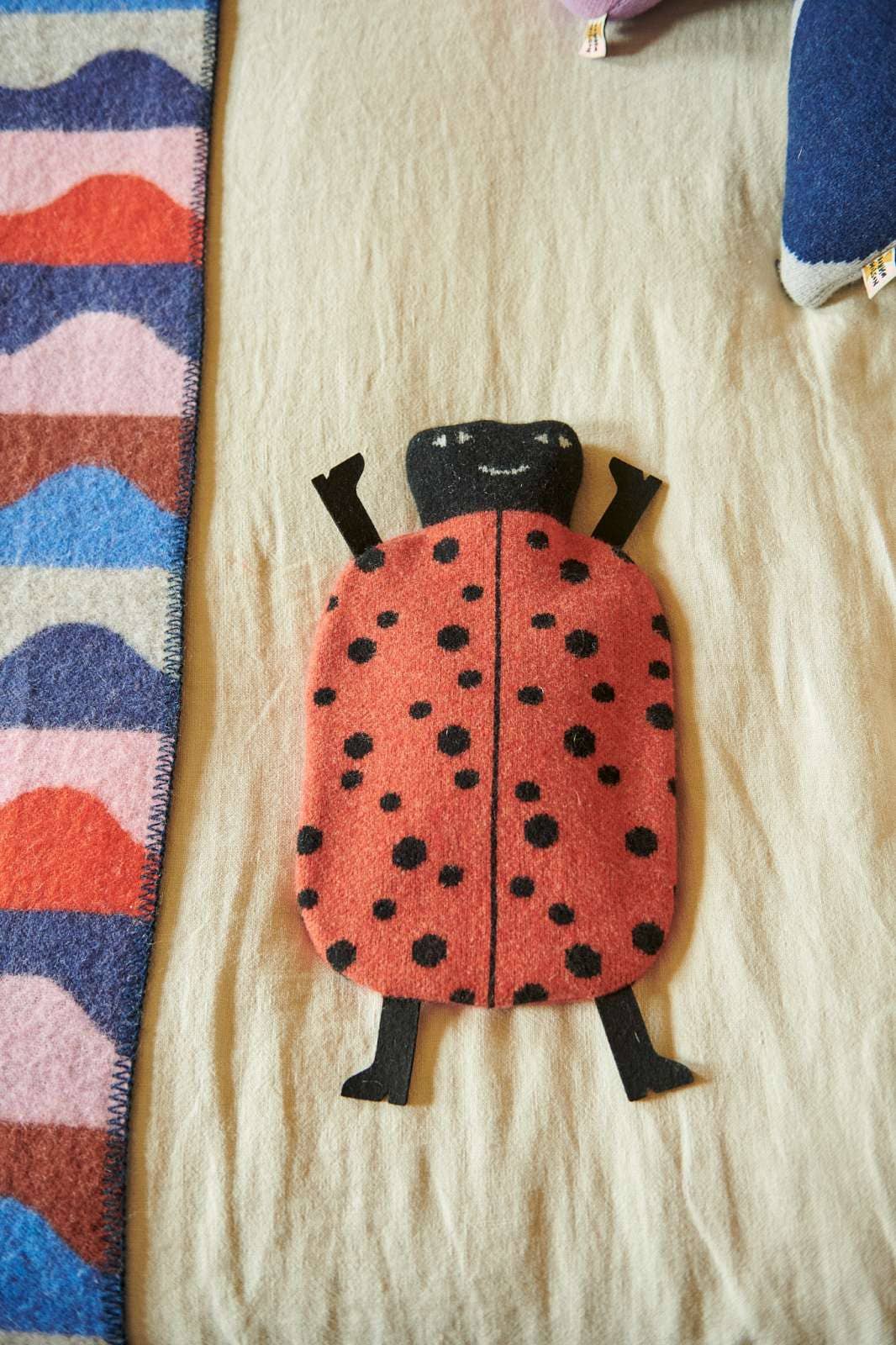 A ladybug-shaped hot water bottle cover made from red-orange felt with black spots and black legs, placed on a beige fabric surface next to a patterned textile strip.