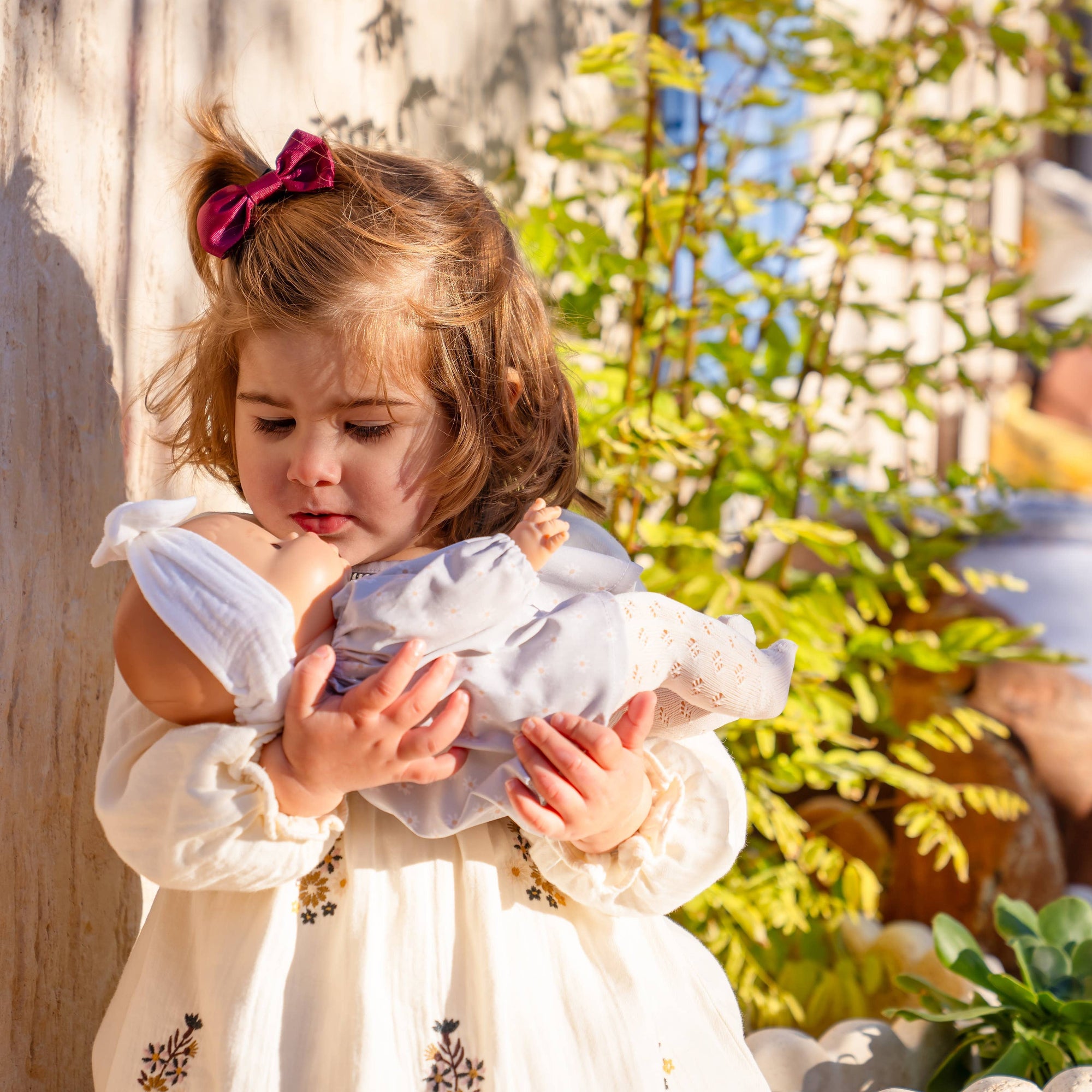 Young girl holding a baby dressed in white outdoors with plants in the background