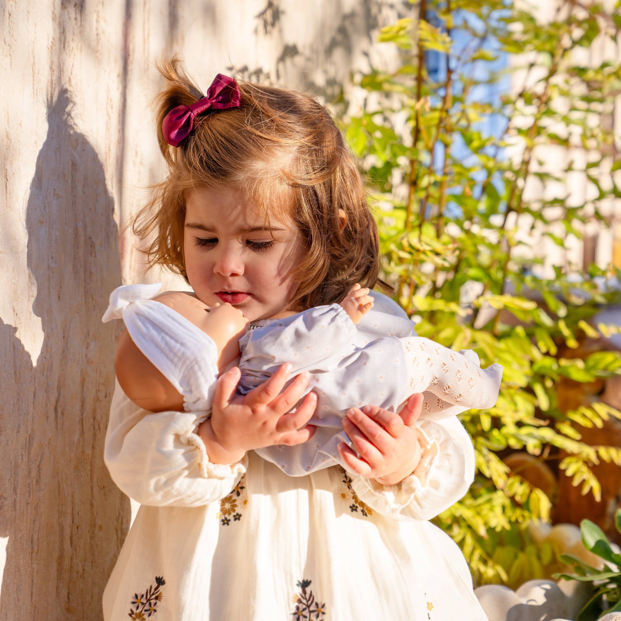 Young girl holding a baby in a garden setting with plants and sunlight.