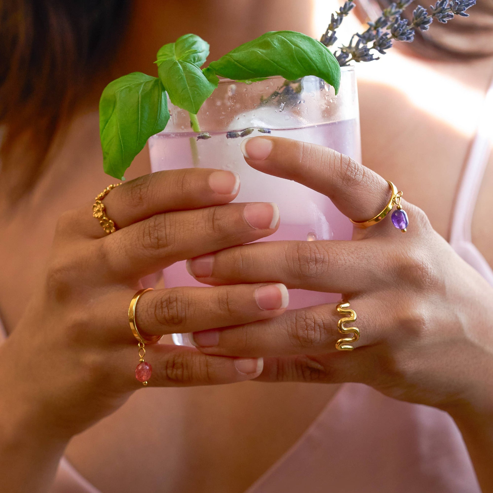 Person holding a glass with a plant and lavender, wearing gold rings.