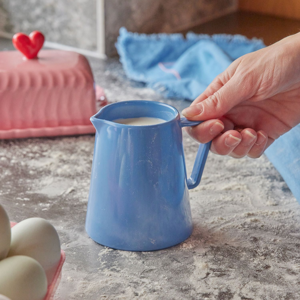 Person pouring milk from a blue pitcher on a kitchen counter with eggs and a cake in the background.