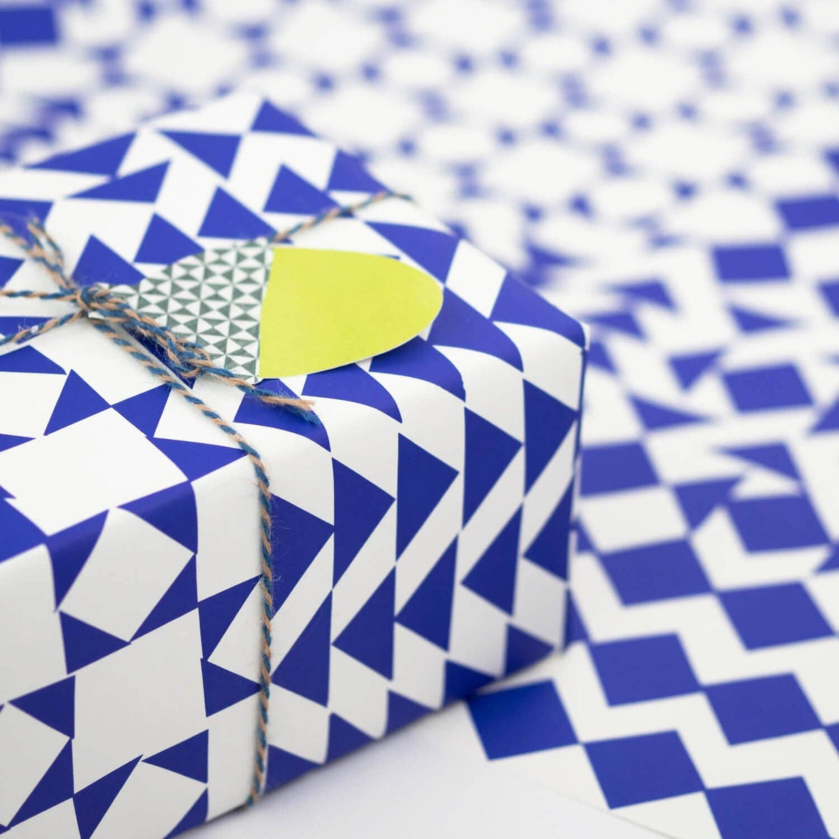 A gift box wrapped in blue and white geometric patterned paper, tied with twine and topped with a small yellow tag, set against a matching patterned background.