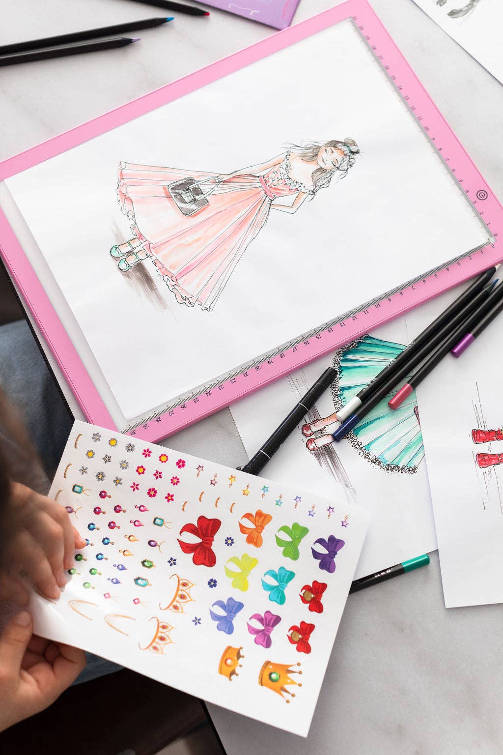 Colorful hand-drawn designs on a white sheet with a pink border, surrounded by art supplies.