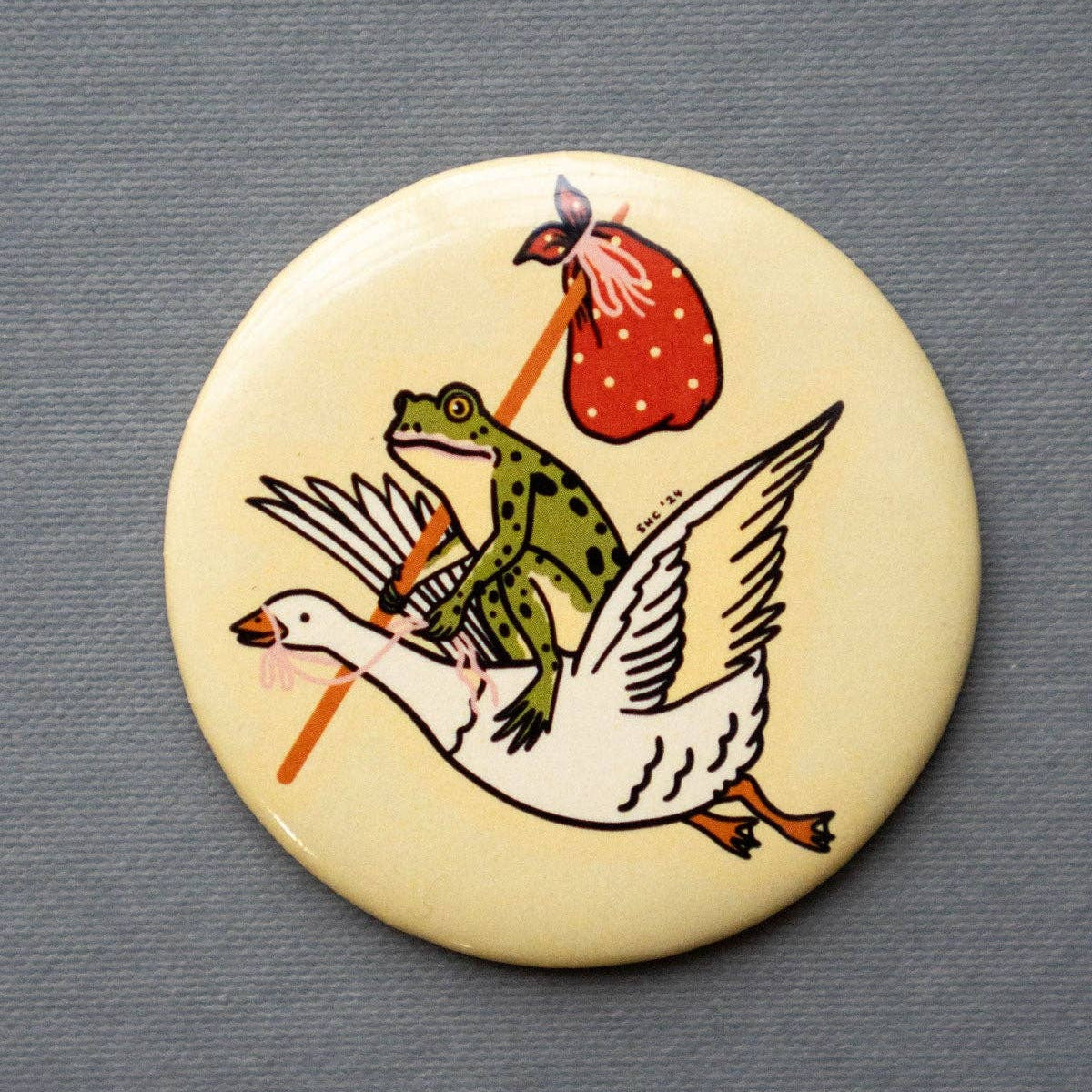 Button badge with a design of a goose carrying a frog on its back, all on a beige background.