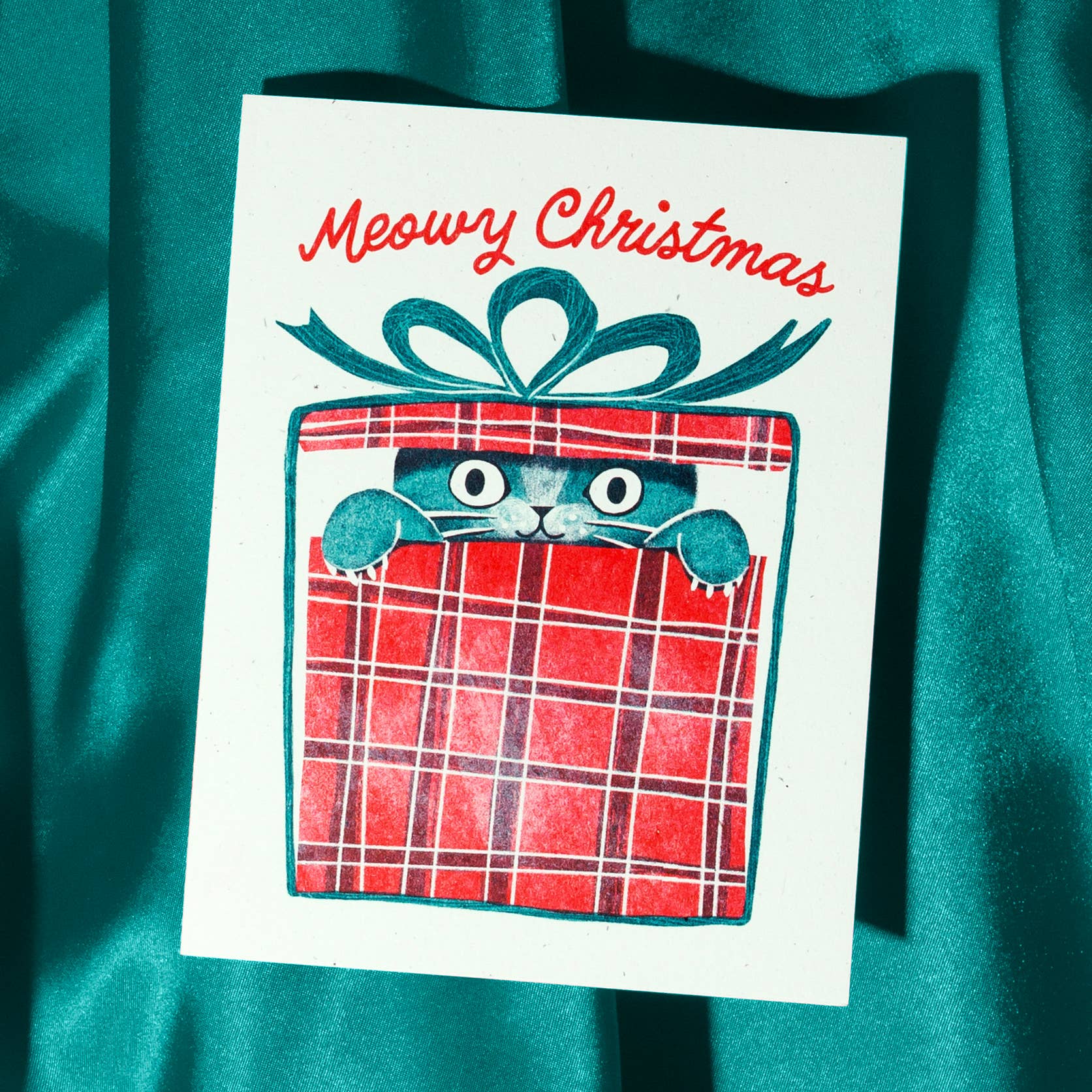 A white Christmas greeting card featuring a cute illustration of a cat peeking out from a red plaid gift box with a teal bow, with the text 'Meowy Christmas' in red script. The card is photographed on a shiny teal fabric background.