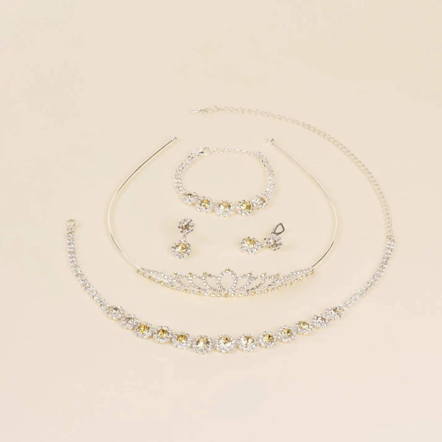 A coordinated jewelry set on a warm cream background, including a silver-tone tiara necklace, matching bracelet, earrings, and a curved collar-style necklace with clear rhinestones.
