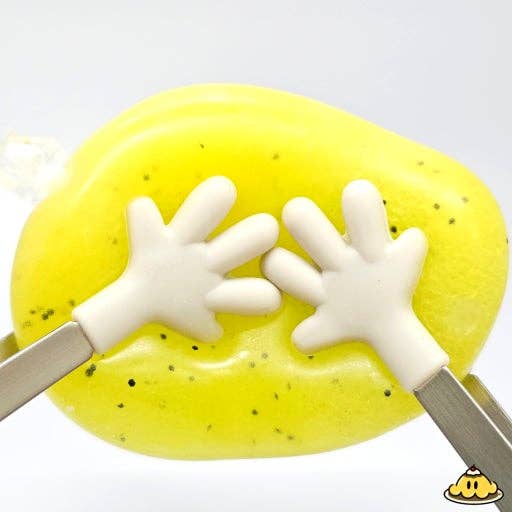 A close-up of a bright yellow slime or clay blob with small black specks, held from both sides by white hand-shaped tools with metallic handles, on a white background.