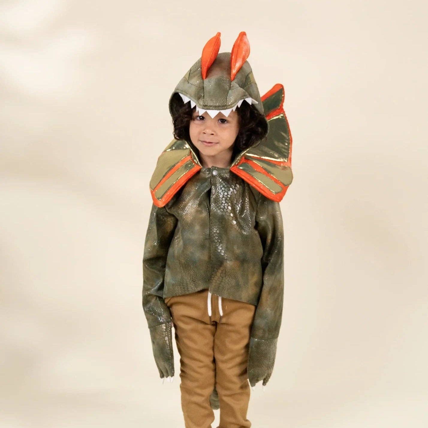 A child wearing a handmade monster or dragon hooded jacket with orange spikes and a camouflage pattern, paired with brown pants and beige sneakers, standing against a plain beige background.