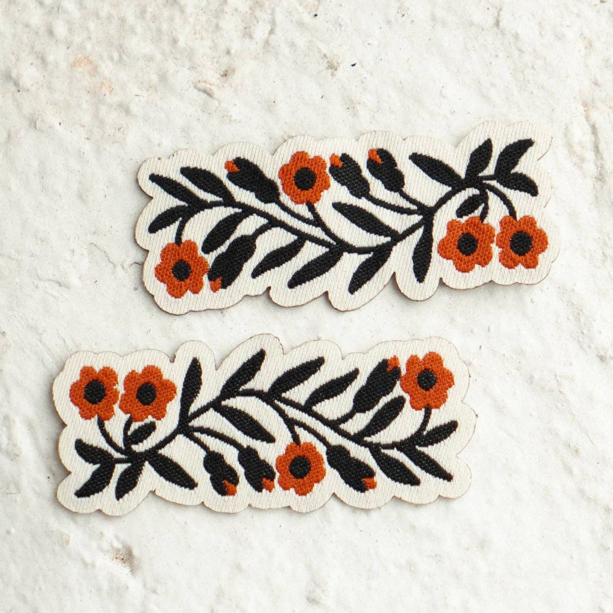 Two matching embroidered floral patches with black leaves and orange flowers on a white fabric backing, displayed on a textured off-white surface.