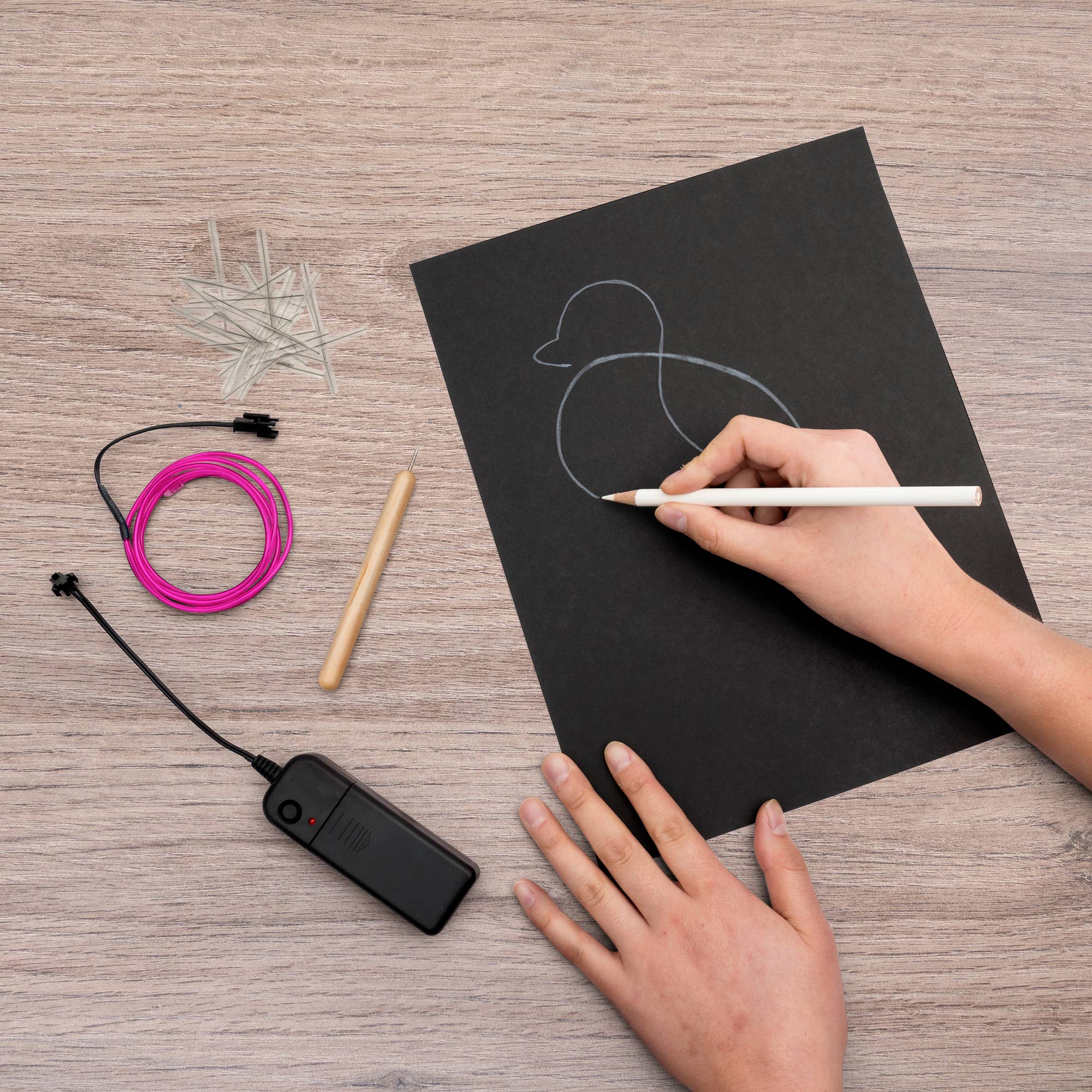 Person drawing a duck on a black sheet of paper with electronic components on a wooden surface.