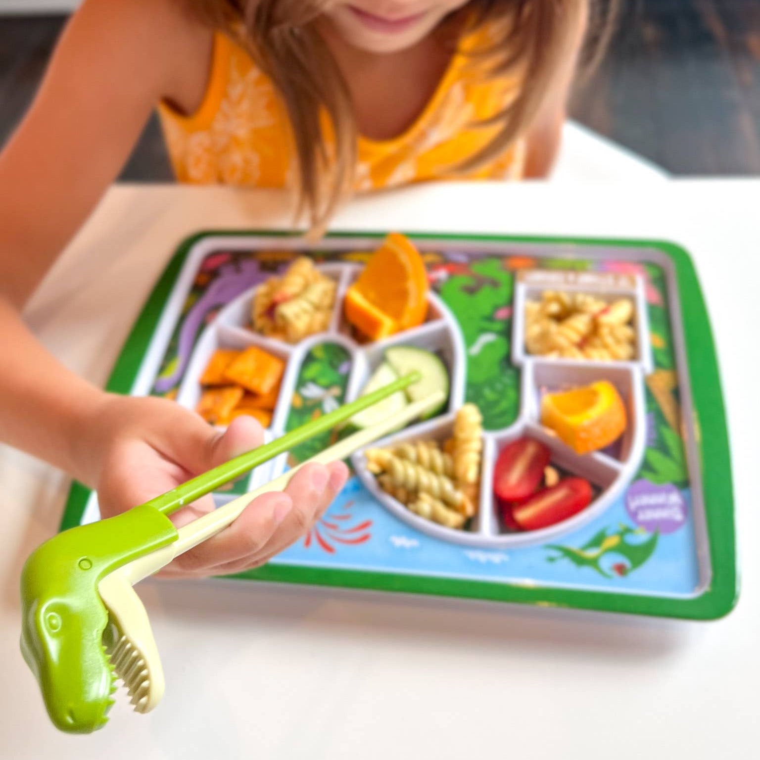 Child using a green dinosaur-shaped fork to eat from a tray with compartments of food.