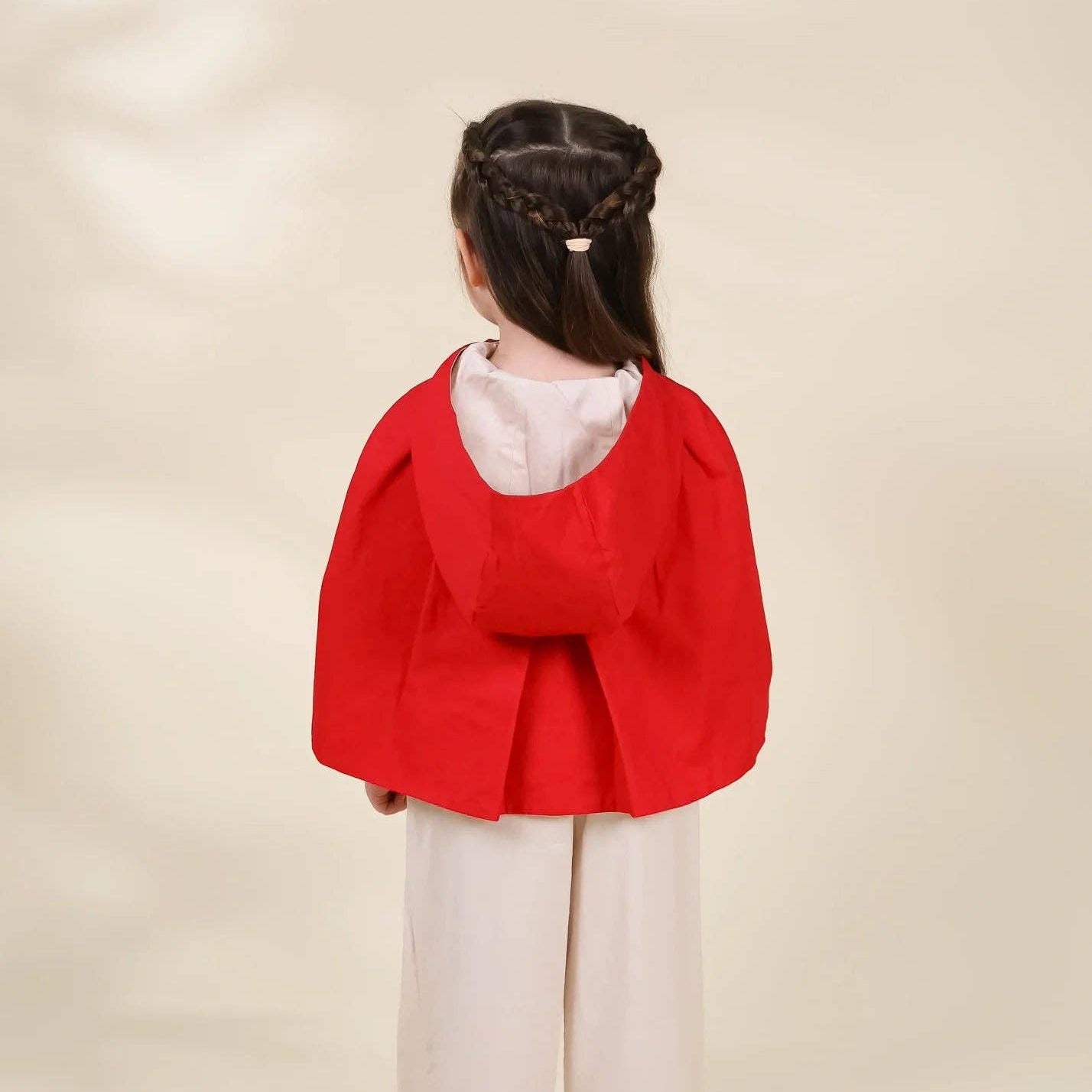 A young girl viewed from behind wearing a bright red hooded cape-style jacket over a white top and white wide-leg pants, standing against a plain warm beige studio background.
