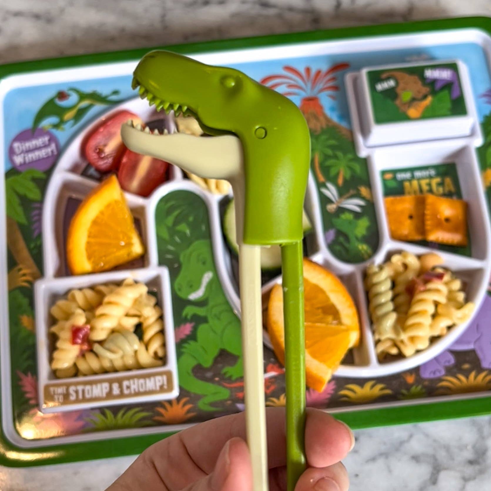 Green dinosaur-shaped fork held over a children's lunchbox with food and dinosaur design.