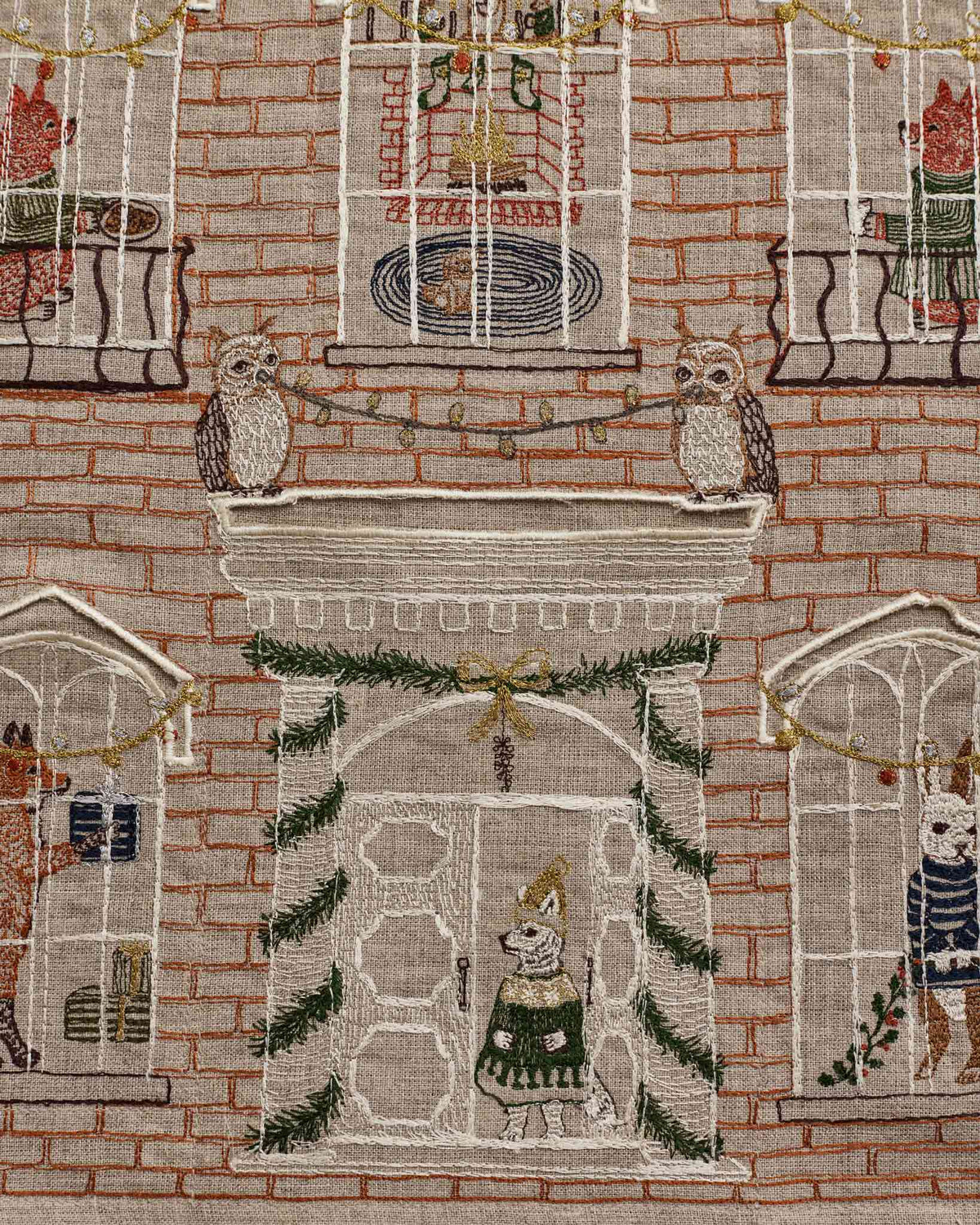 A close-up of a woven textile featuring a brick building facade with arched windows, owl figures on mantels, holiday garlands, and small illustrated characters in sweater vests.