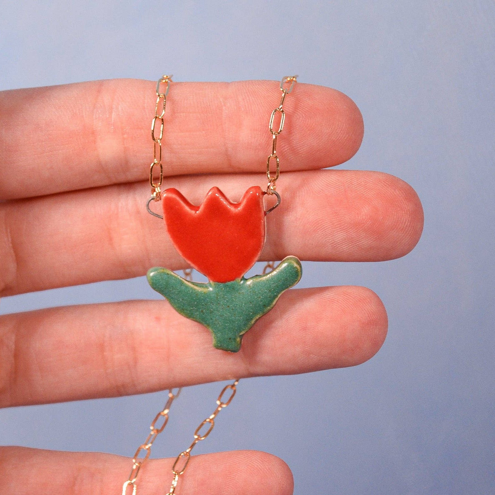 Hand holding a necklace with a red tulip pendant against a blue background