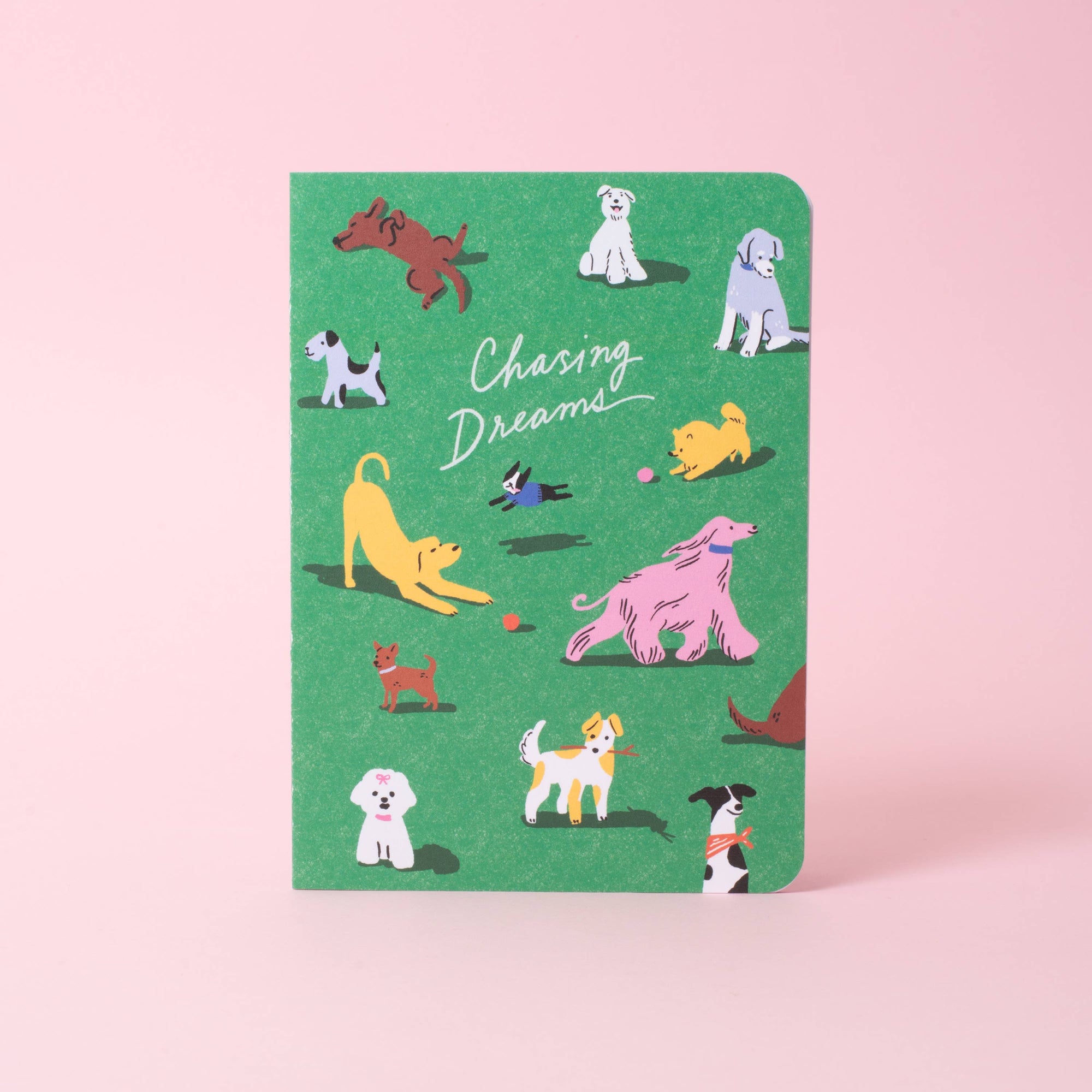 A green notebook with a playful illustration of various dog breeds and the text 'Chasing Dreams' on the cover, set against a solid pink background.