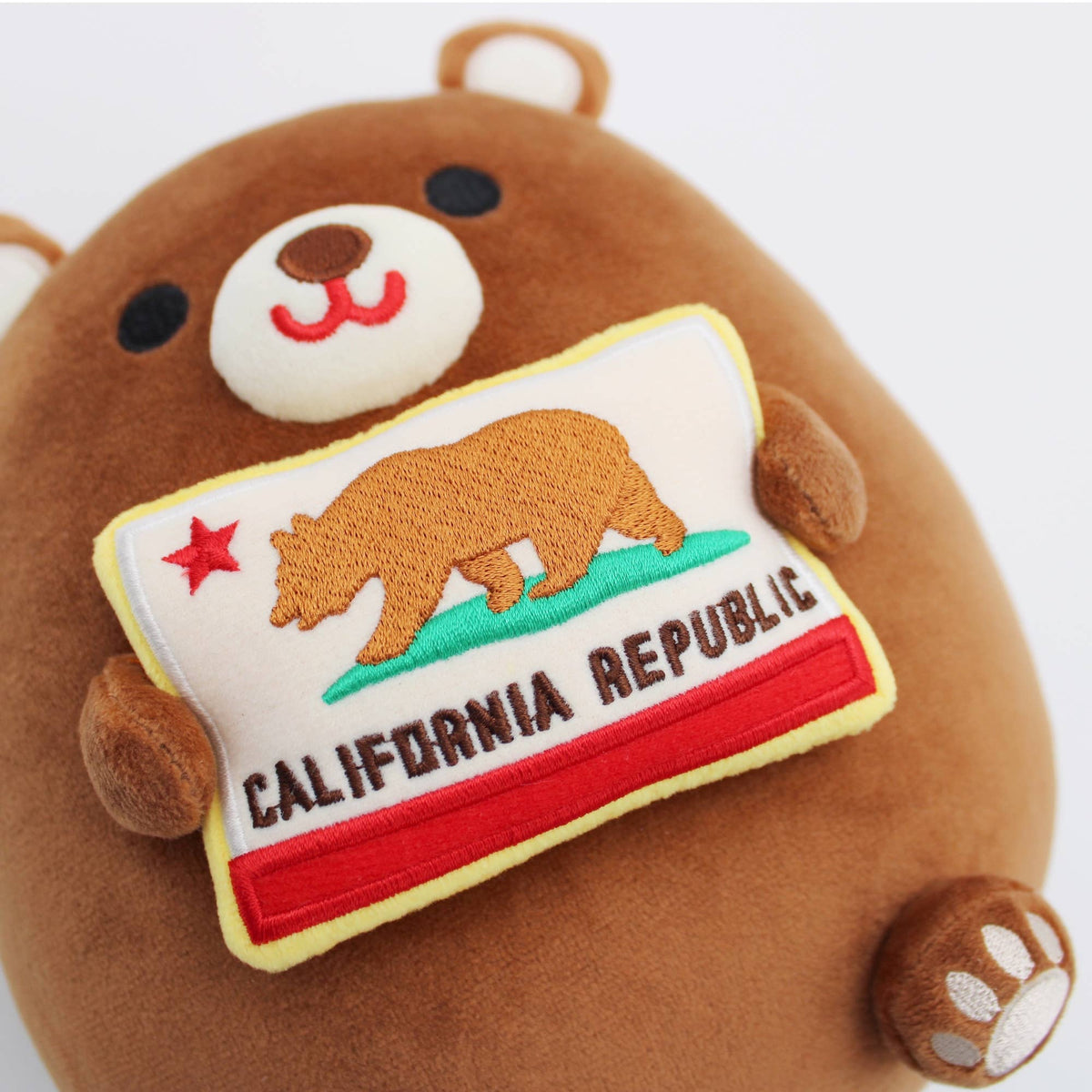 A close-up of a brown plush bear holding a California Republic flag patch. The plush has a cream snout, black eyes, a red stitched smile, and a paw print on its foot.