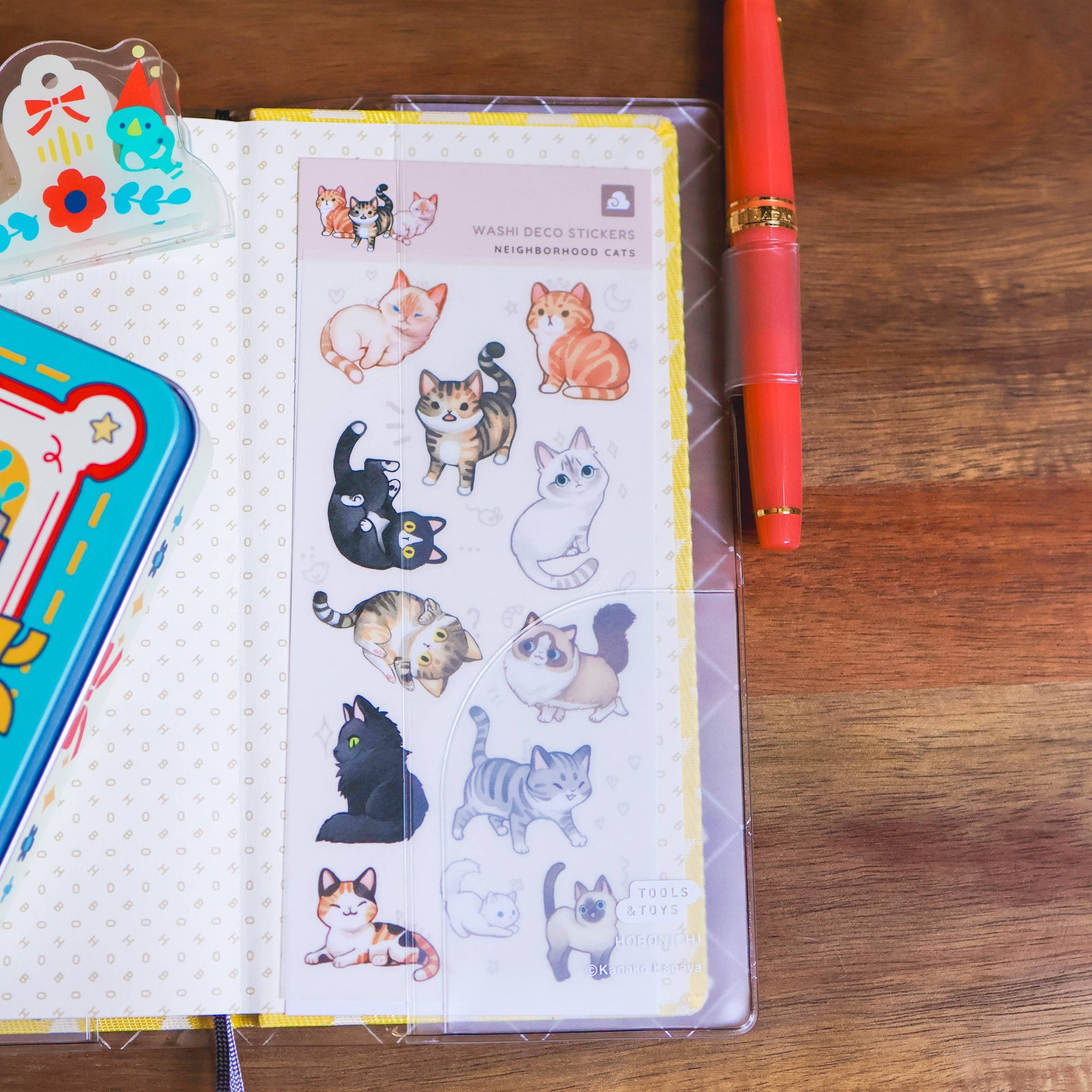 Sheet of cat stickers in a clear plastic case on a wooden surface with a red pen.