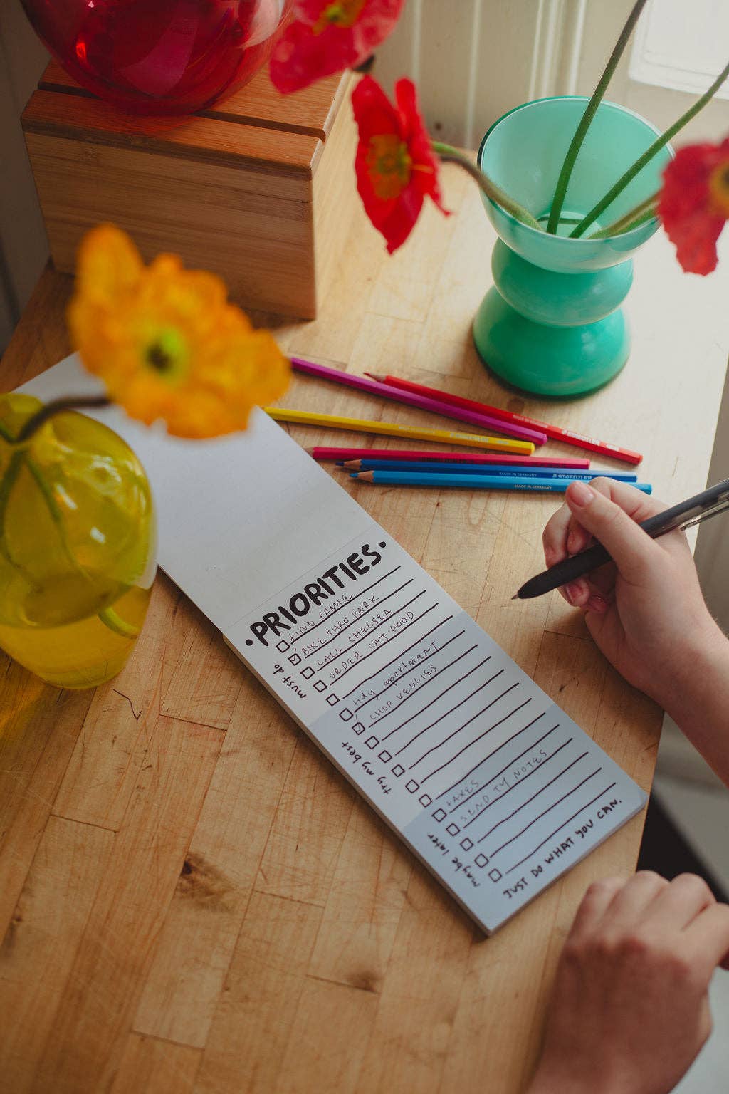 Person writing on a 'Priorities' list with colorful pencils and flowers on a wooden table.