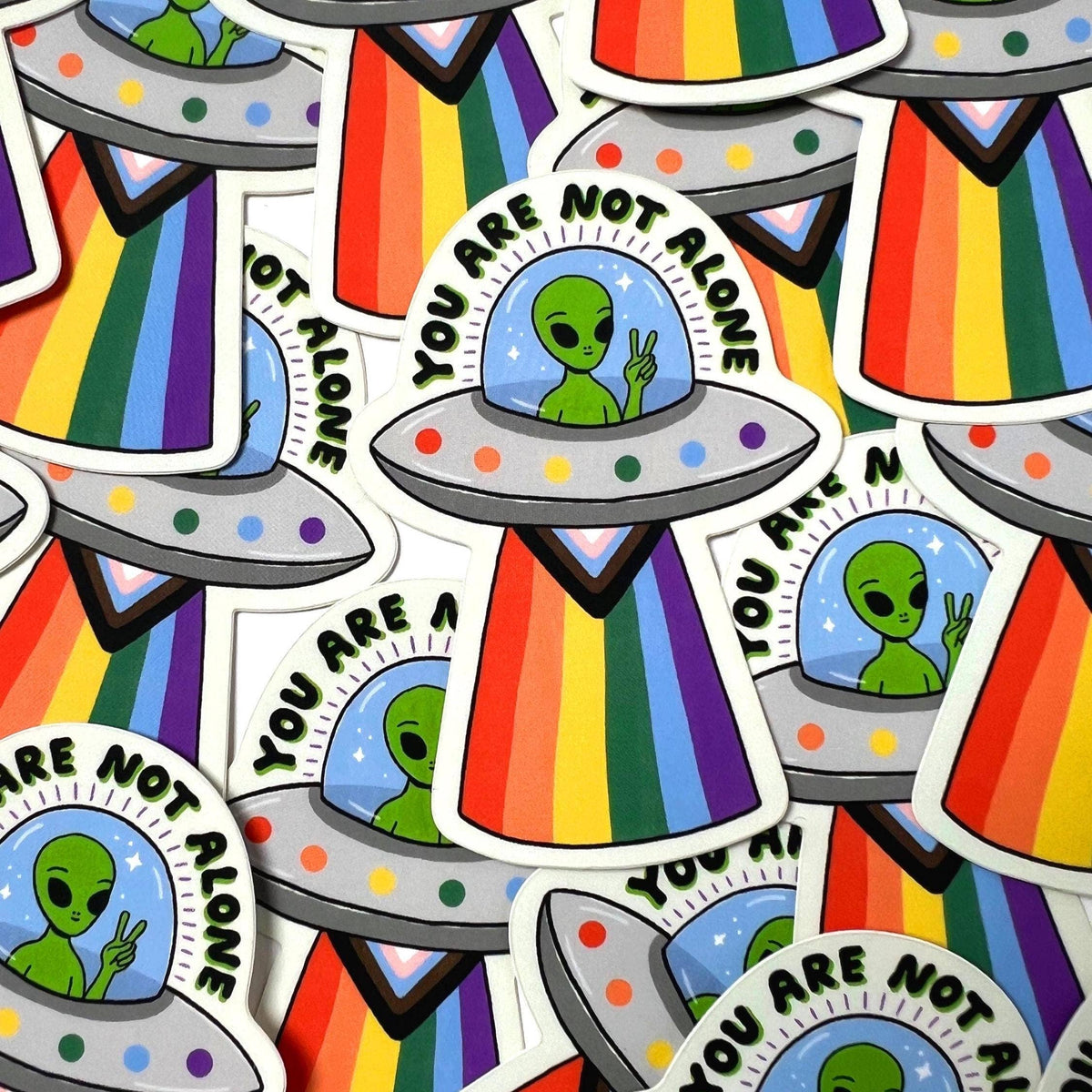 A pile of die-cut stickers featuring a green alien in a UFO with rainbow stripes, with one sticker clearly showing the text 'YOU ARE NOT ALONE'.