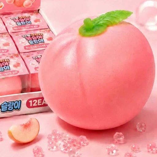 A pink peach-shaped squishy toy with a green leaf on top, shown next to its pink retail packaging box and scattered pink crystal-like pieces on a pink surface.